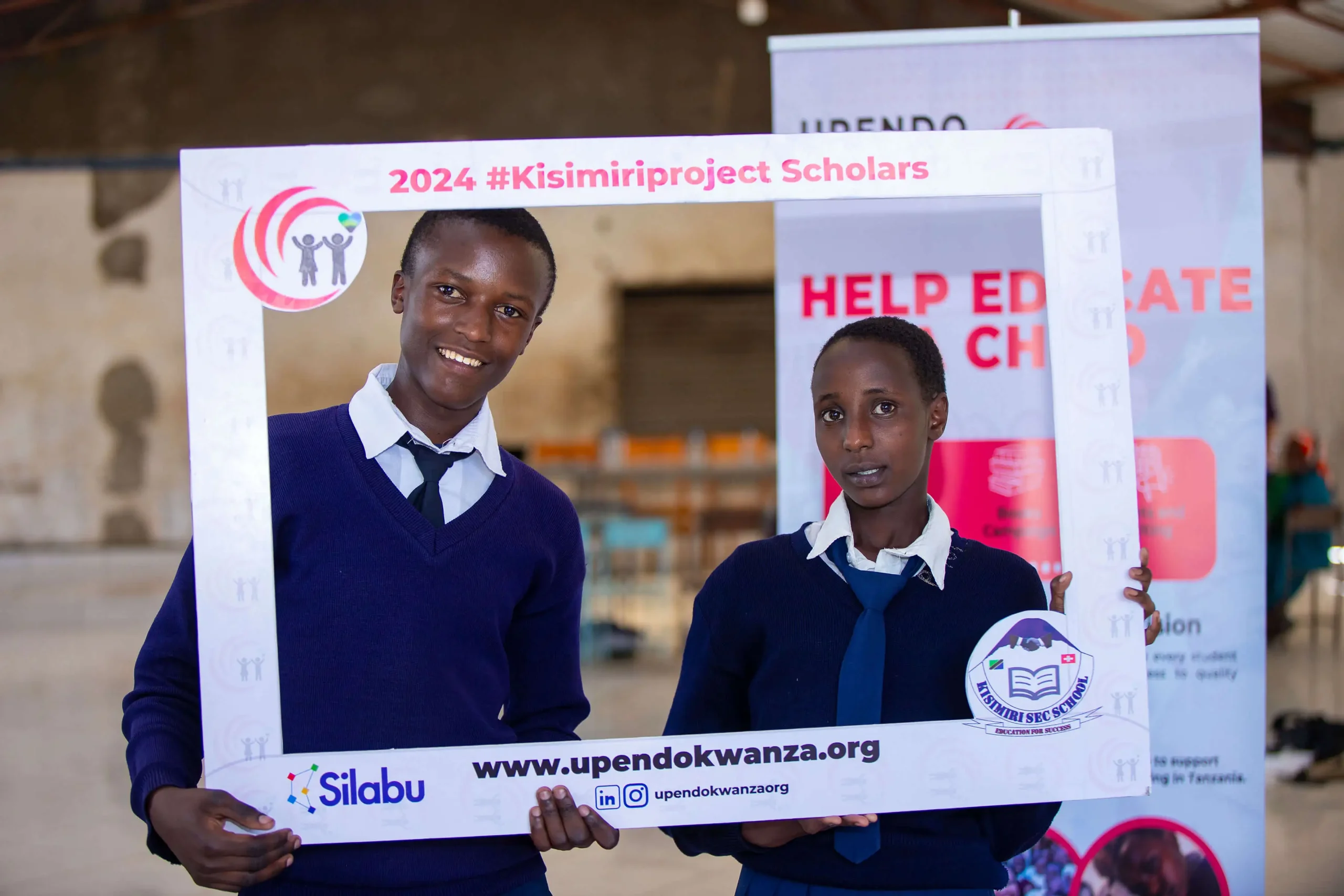 Two young students in blue school uniforms holding a frame sign that reads '2024 Kisimiri Project Scholars' made by the Upendo Kwanza organization