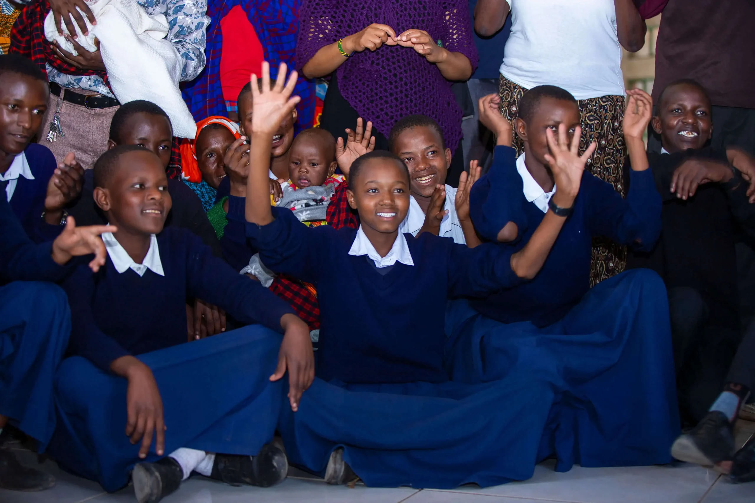 Students sit on the school ground for a group picture, smiling and making hand gestures