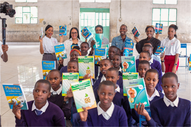 Students sitting in a classroom with Upendo Kwanza volunteers standing in the back, all holding up new textbooks.