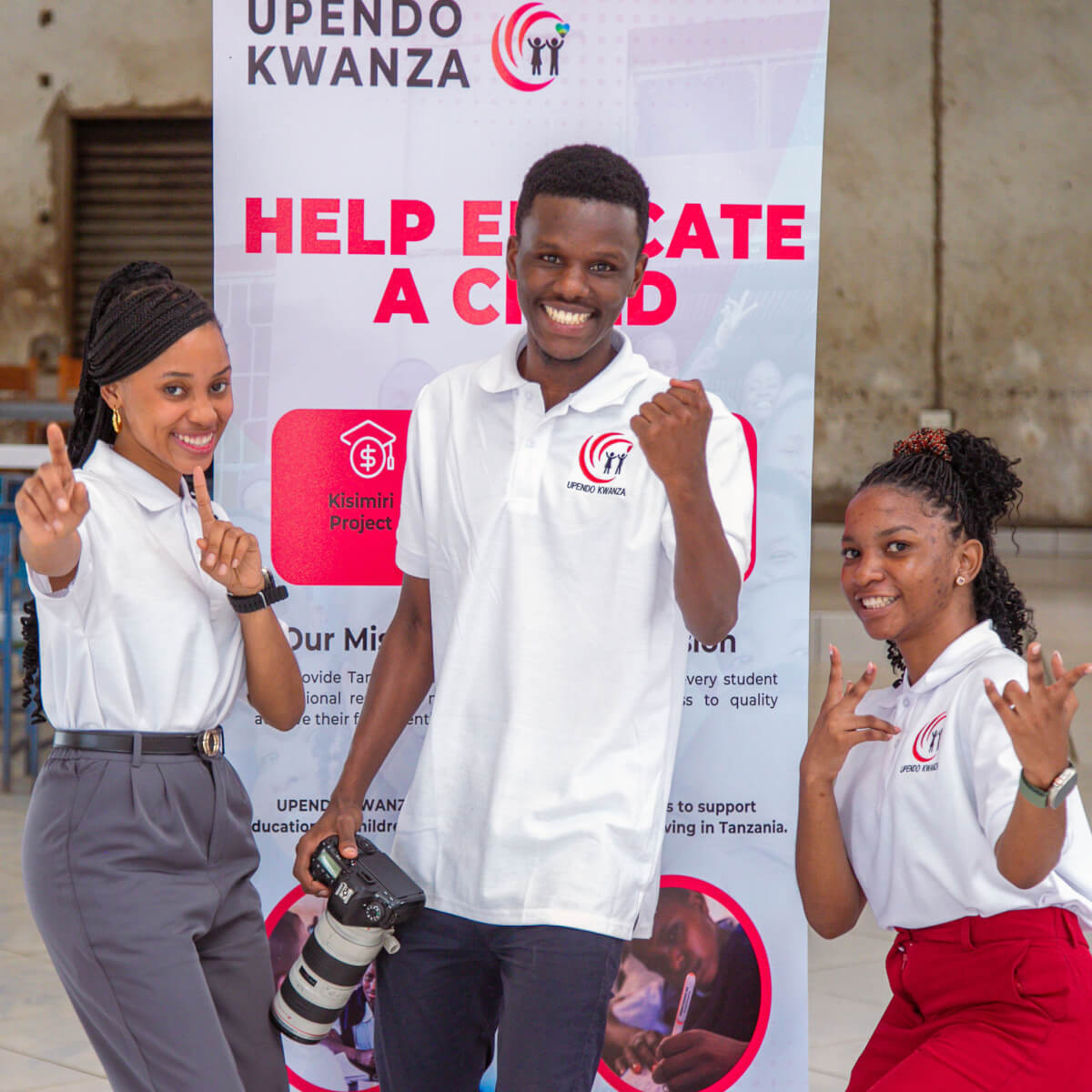 Three volunteers wearing white t-shirts with the Upendo Kwanza logo smile and make hand gestures for the camera