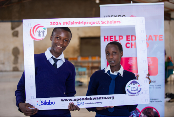 Two young students in blue school uniforms pose for a picture, holding a sign that reads '2024 Kisimiri Project Scholars' made by the Upendo Kwanza organization